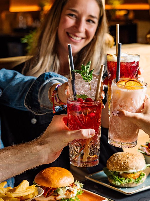2 Frauen stoßen mit je 1 Glas mit 2 weiteren Personen, von denen nur die Hände mit je 1 Glas zu sehen ist an. Sie lächeln. Pommes und Burger liegen auf einem Teller vor den 4 Personen.
