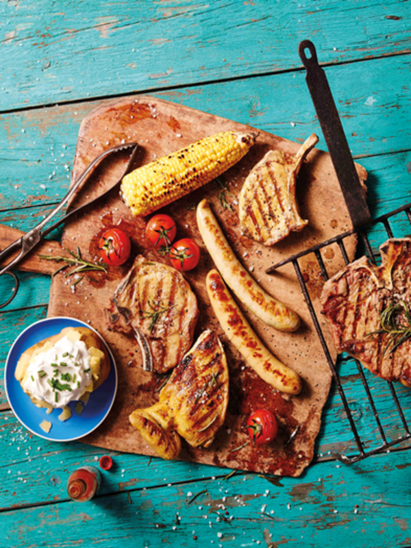 Blauer abgenutzter Holzboden, darauf ein Holbrett mit Grillfleisch, Tomaten, Mais, Würstchen und Ofenkartoffel