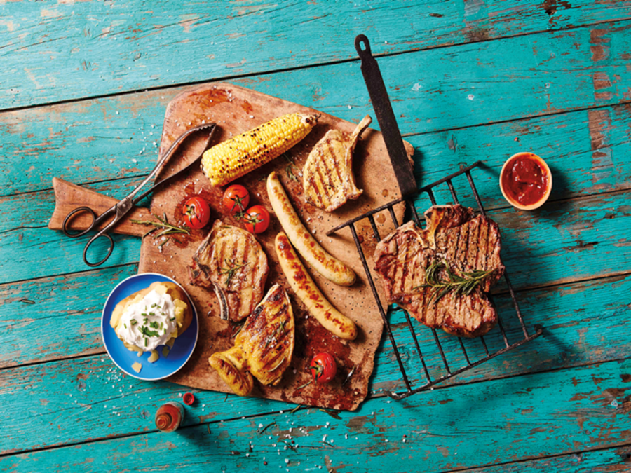 Blauer abgenutzter Holzboden, darauf ein Holbrett mit Grillfleisch, Tomaten, Mais, Würstchen und Ofenkartoffel
