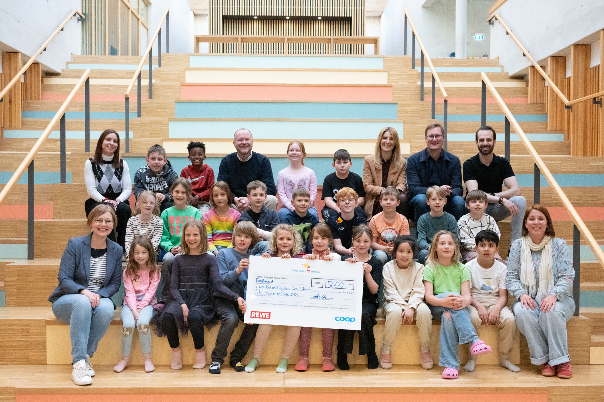 Gruppenfoto von Kindern und Erwachsenen auf einer großen Holztreppe in einem hellen Innenraum. In der Mitte halten mehrere Kinder einen großen Spendenscheck über 5.000 Euro mit REWE- und coop-Logo in die Kamera.