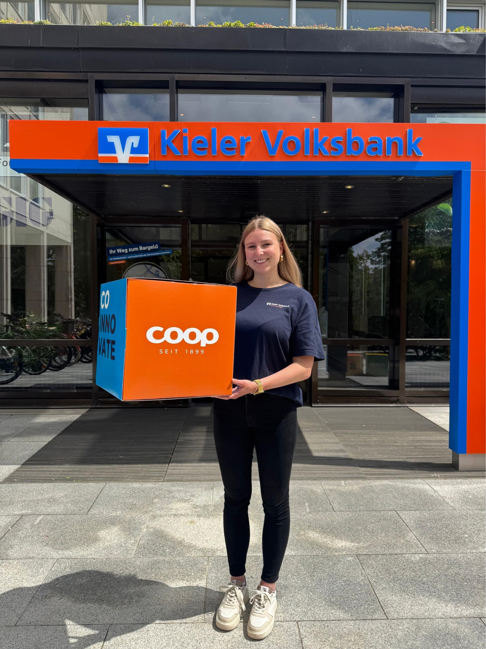Blonde junge Frau hält einen orange farbenen Würfel mit dem weißen coop Logo in die Kamere. Hinter ihr ist der Eingang in eine Kieler Volksbank, was man an dem Logo und großen Schriftzug über ihr erkennnt.
