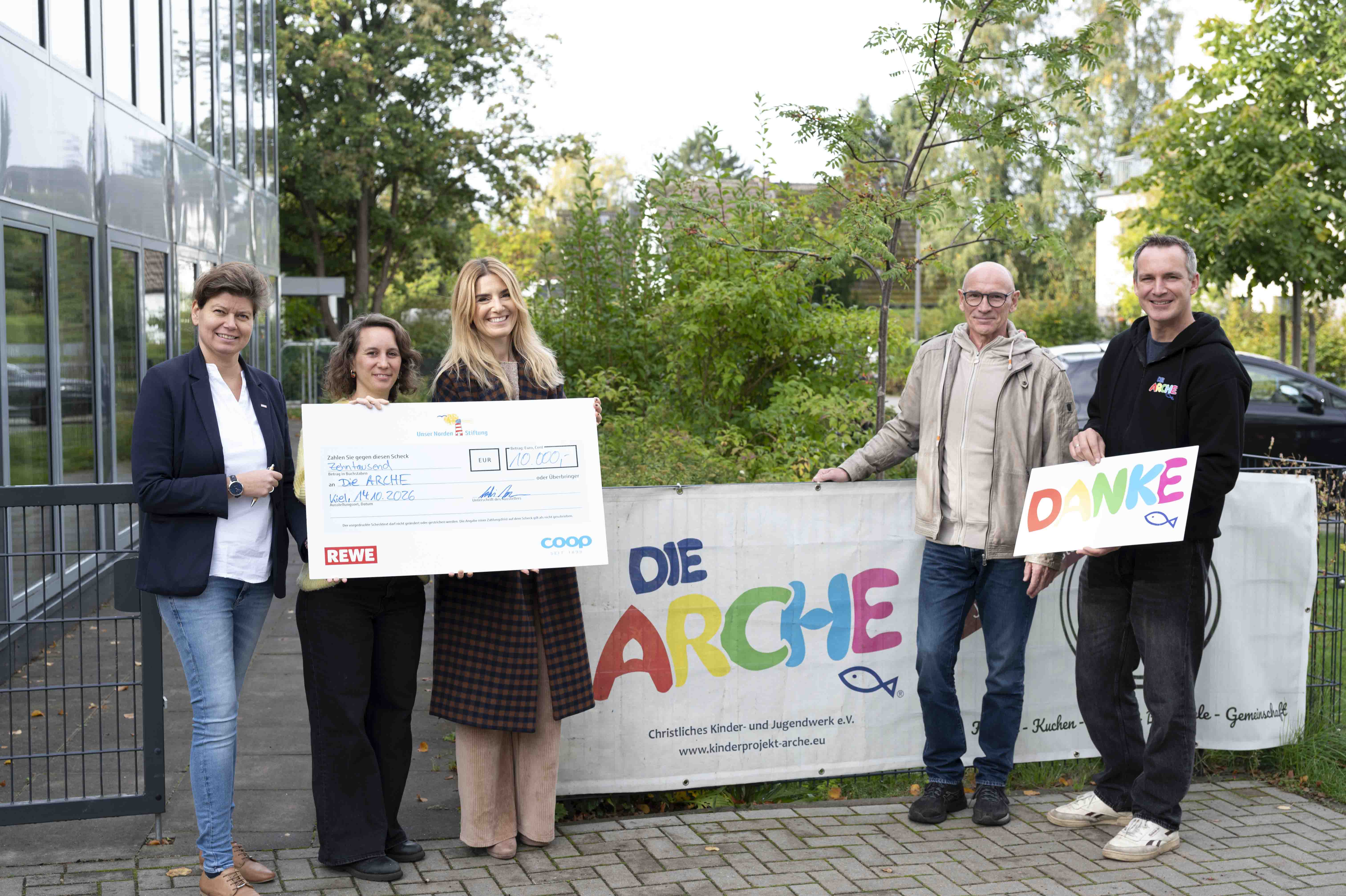 Mitarbeiterinnen und Mitarbeiter von der coop, REWE und Arche halten einen offiziellen Spendencheck im Wert von 10.000 Euro in die Kamera. 