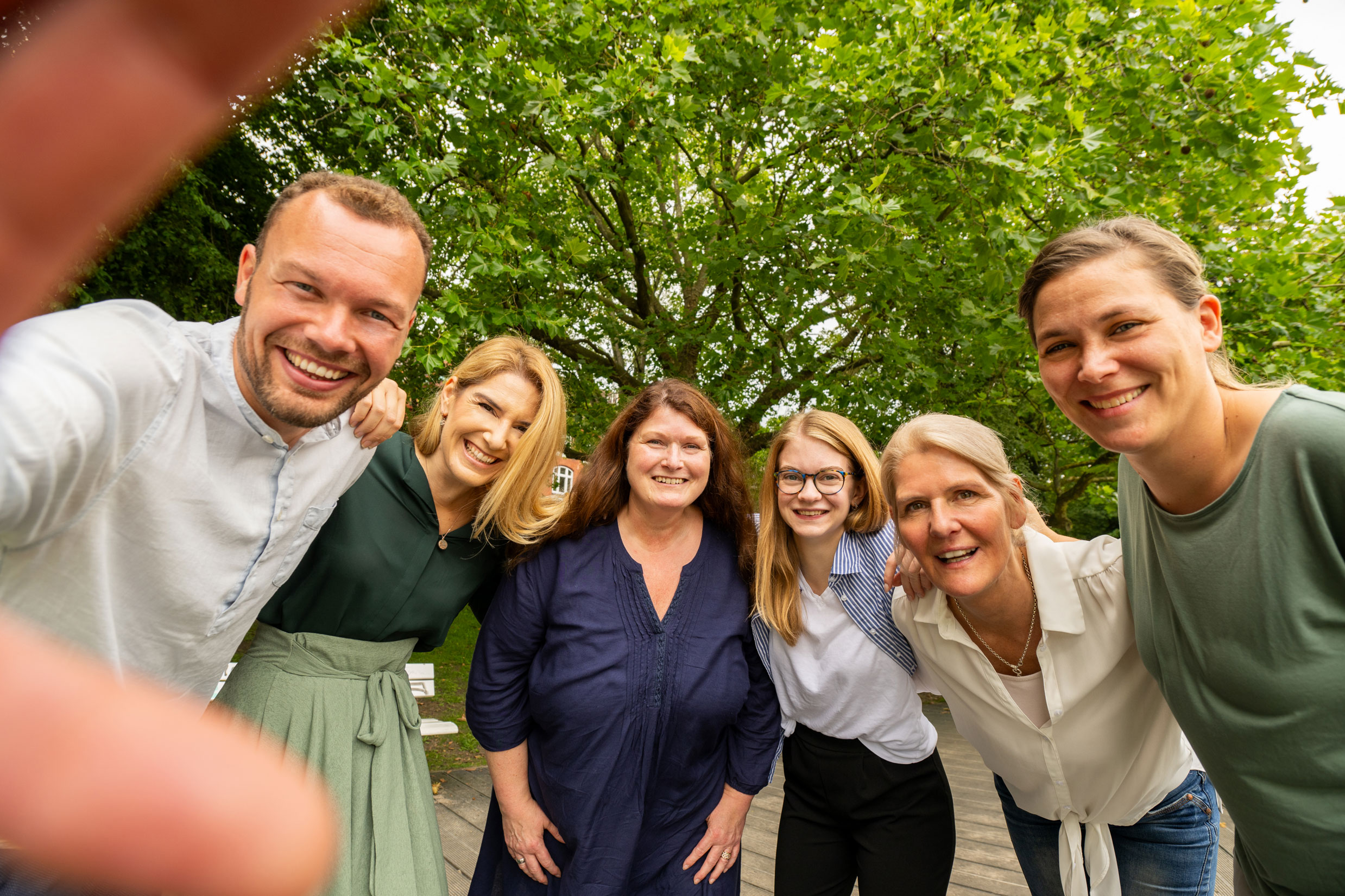 Ein junger Mann macht von sich und fünf Frauen im Halbkreis ein Selfie. Alle schauen direkt in die Kamera. Man sieht noch zwei Finger des jungen Mannes im Anschnitt. Im Hintergrund sind gründe Bäume.