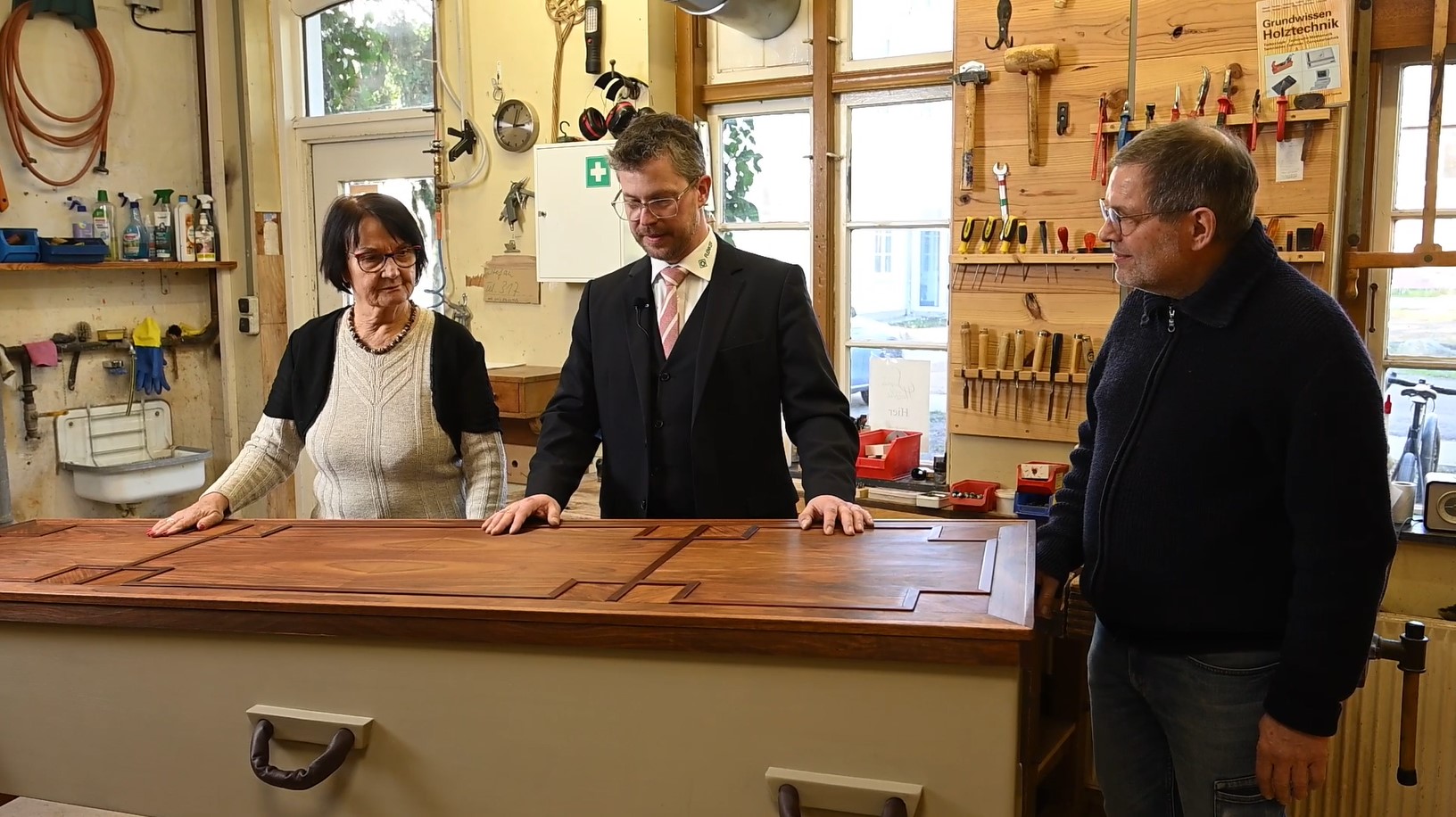 Eine Frau steht an der langen Seite eines geschlossenen Sarges 1 Hand auf dem Sarg, rechts daneben steht ein Herr und hält beide Hände auf den Sarg. An der Stirn des Sarges, auf der rechten Seite steht ein weiterer Herr. Es scheint sich um Mitarbeiter des Unternehmens zu handeln.