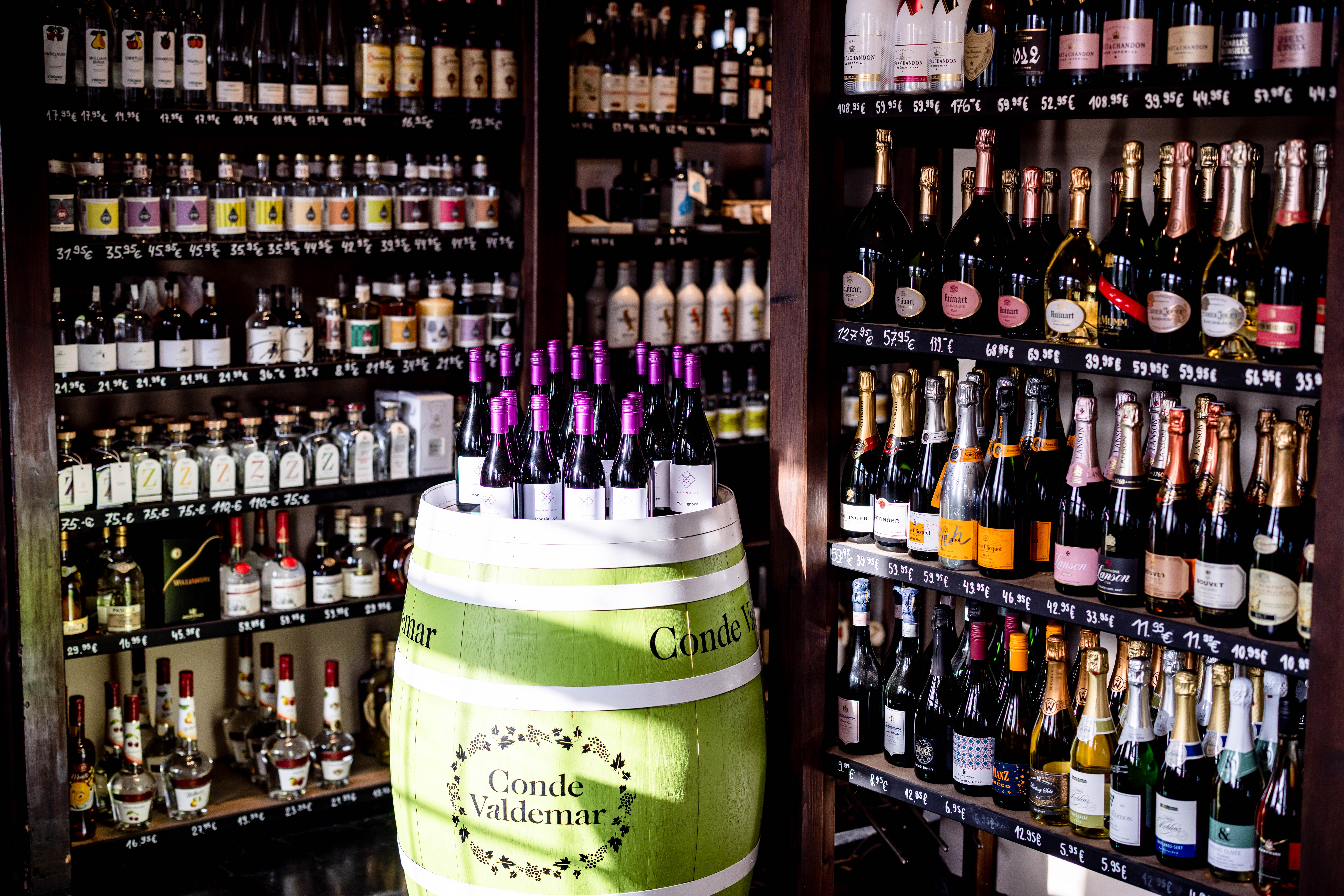 Ein über Eck gefülltes Weinregal Deckenhoch. Davor steht ein grün/weißes Weinholzfass mit vielen Flsschen Wein einer gleichen Sorte