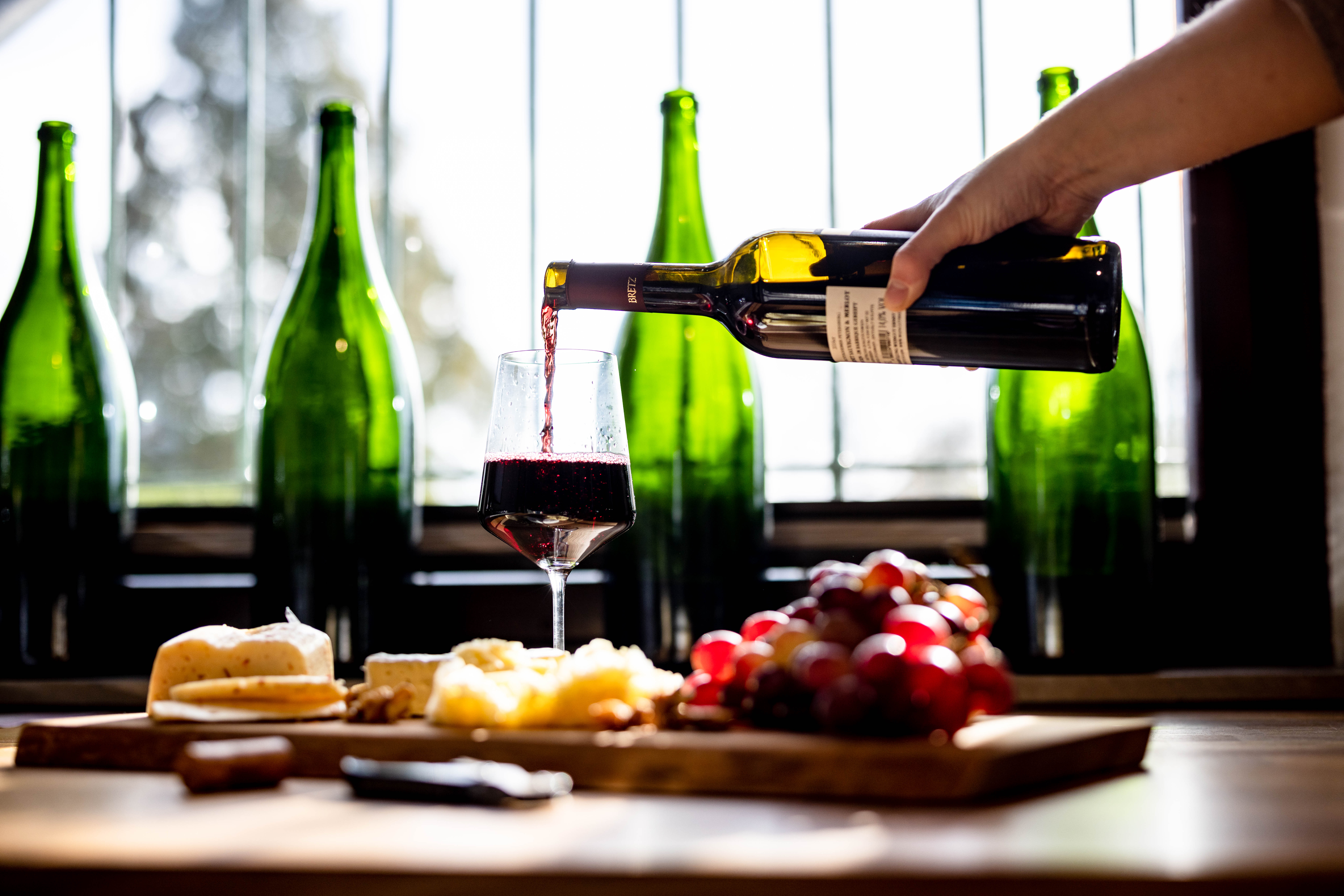 4 grüne Flaschen Wein im Hintergrund. Im Vordergrund ein Weinglas zur Hälfte mit Rotwein gefüllt. Eine Hand schenkt derweil noch mit einer Flasche in das Glas ein. Das Glas steht auf einem Holzbrett, welches mit Brot, Weintrauben und Käse bestückt ist