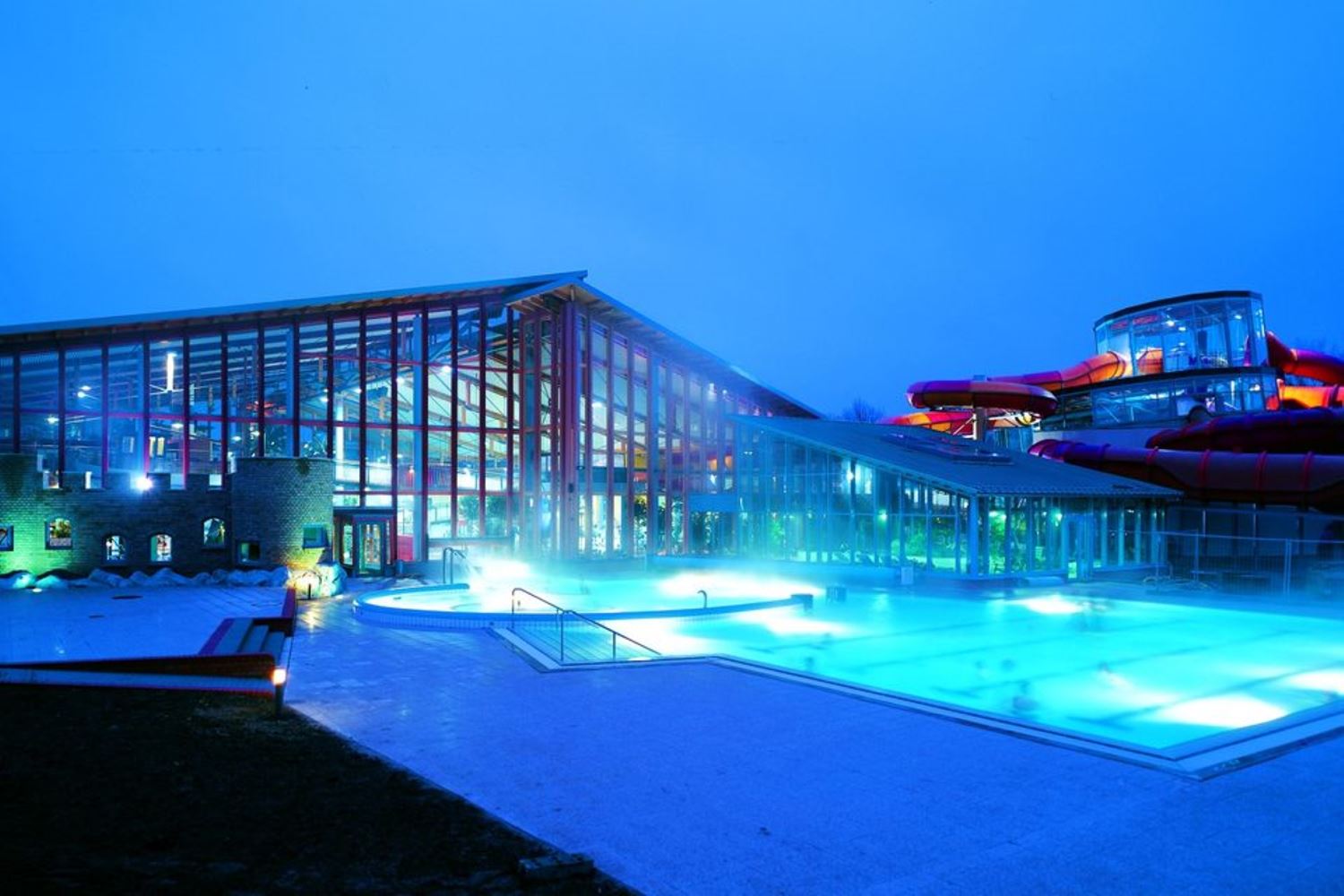 Der Außenpool vom Wonnemar ist im Vordergrund vor dem Gebäude blau beleuchtet. Das Gebäude im Hintergrund hat eine Glasfront und auf der rechten Seite einen Rutschenbereich. Der Himmel ist dunkel, da das Bild abends bei Dämmerung aufgenommen wurde. 