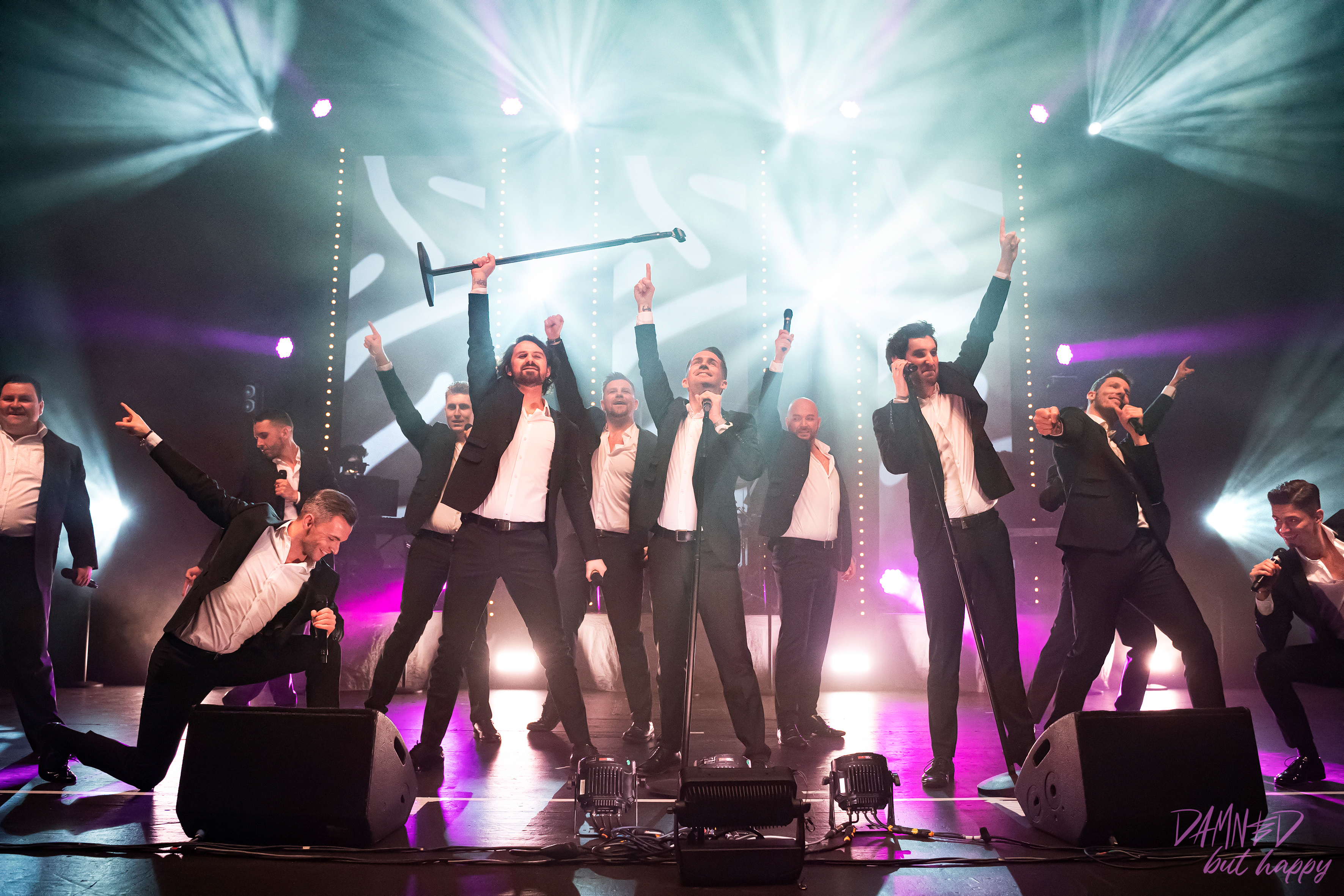 12 Tenöre stehen auf der Bühne und heben jeweils einen Arm in die Luft, darüber ist helles Scheinwerferlicht