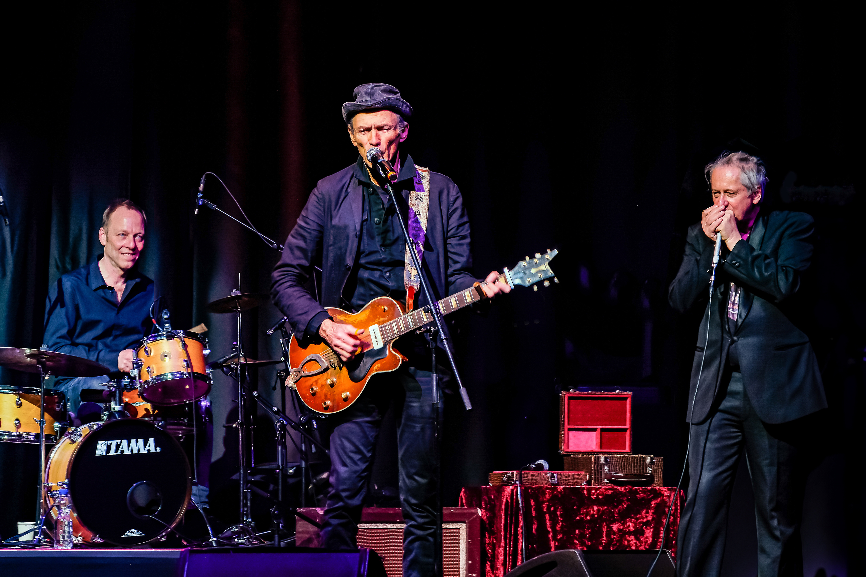 Dunkler Hintergrund, im Vordergrund ein zugleich singender und Gitarre spielender Mann mit Hut, links daneben ein Schlagzeuger, rechts daneben ein Mann der Mundharmonika spielt