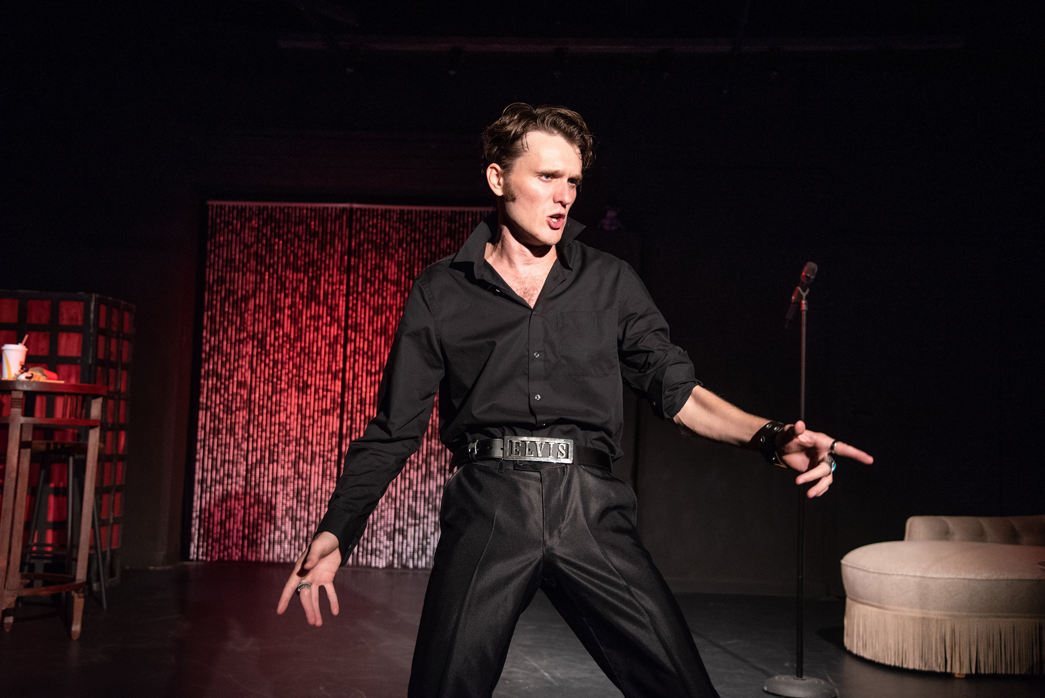 Elvis Darsteller steht in typischer Elvis Pose, Arme etwas ausgebreitet, Finger gespreizt. Er trägt ein schwarzes Hemd und eine schwarze Hose. Im Hintergrund ist ein Lamettavorhang und ein Sofa aus den 60ern