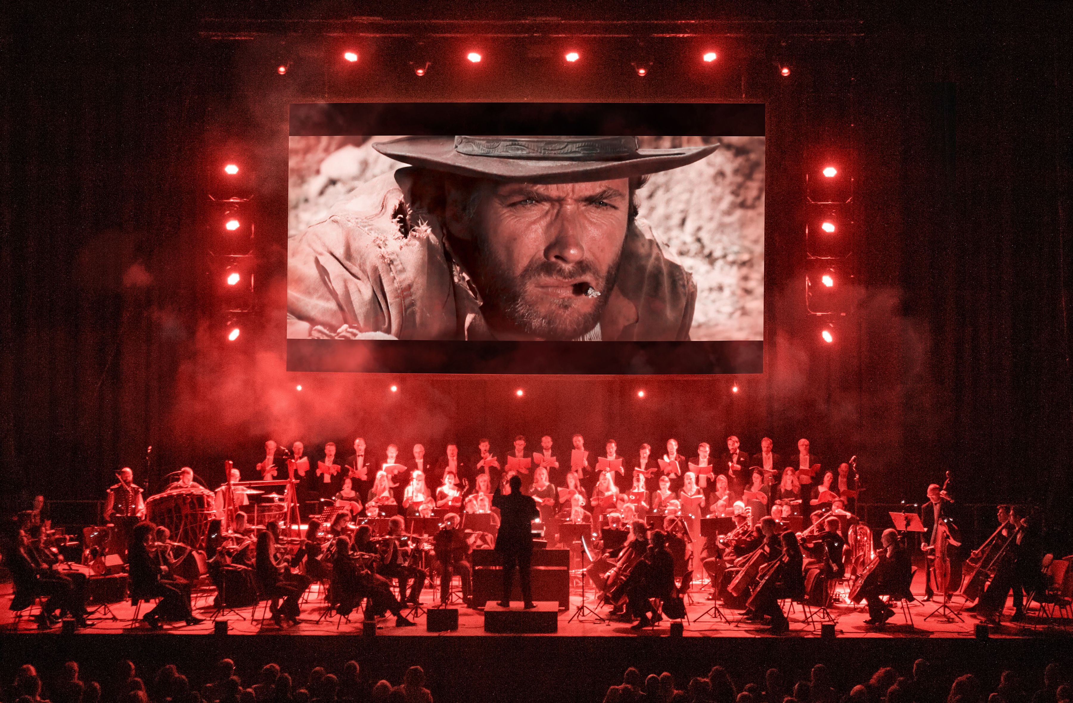 Auf dem Bild ist eine Leinwand mit 3 roten Balken links und rechts und oben. Darunter ist das Gesicht eines Cowboys. Im unteren Bereich ist ein Orchester auf der Bühe, der Dirigent steht in der Mitte