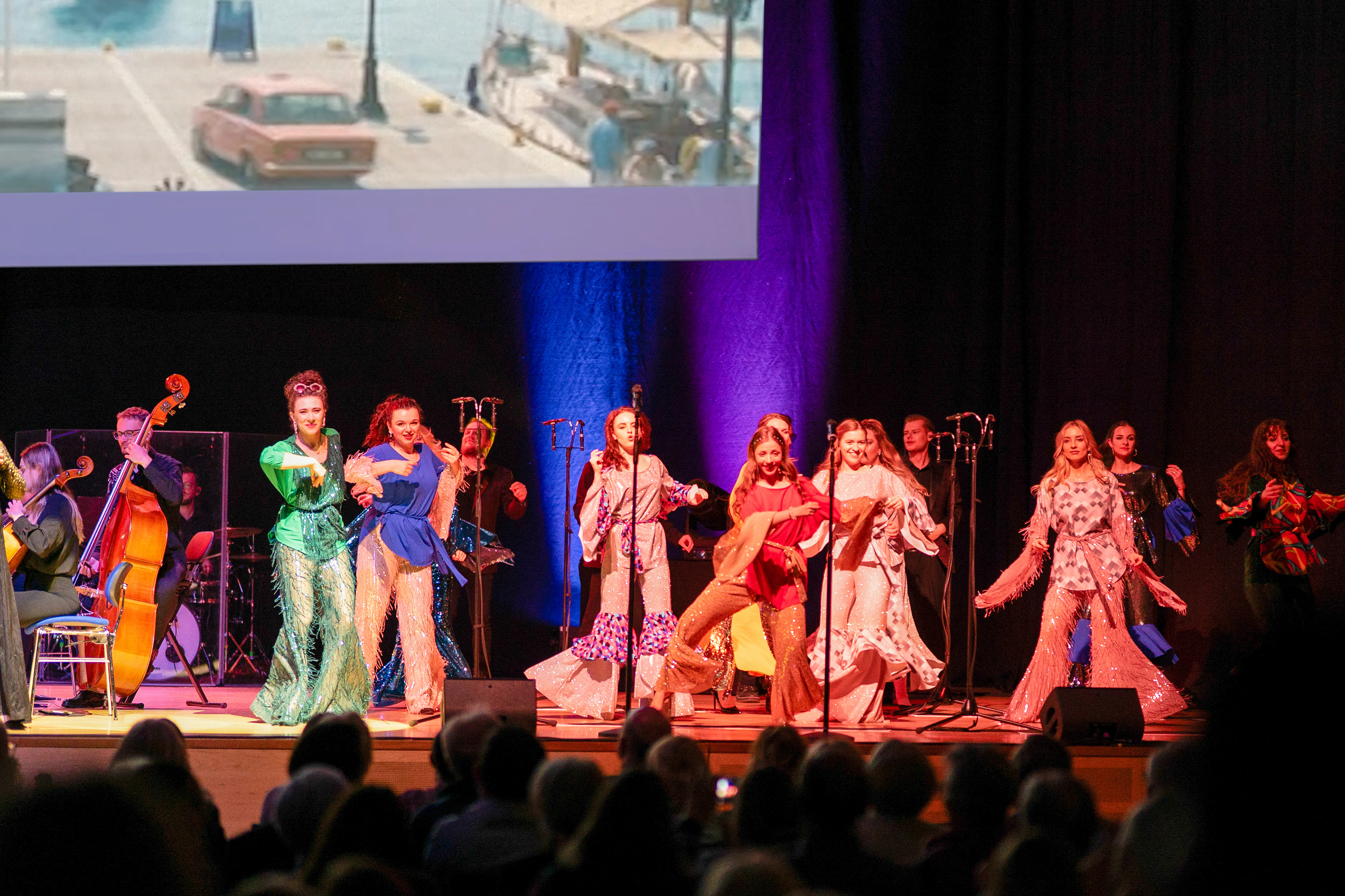 Eine Bühne mit mehreren singenden und tanzenden Frauen in bunten Schlaghosen und Blusen. Darüber ist ein Ausschnitt einer Leinwand. Links im Bild sieht man angeschnitten die Musiker.