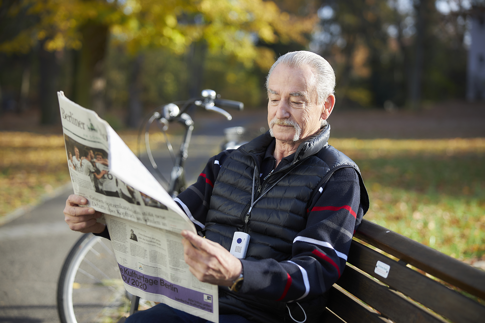 Ein Mann sitzt auf einer Parkbank und liest Zeitung. Im Hintergrund ist sein Fahrrad, sowie eine Wiese und Bäume. 