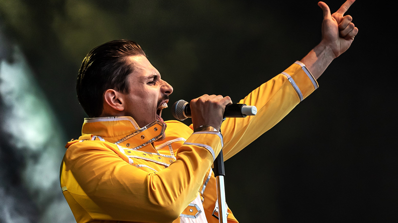 Freddy Mercury Double reißt in einem gelben Jacket den linken Arm in die Luft und hält in der rechten Hand ein Mikrofon an den Mund