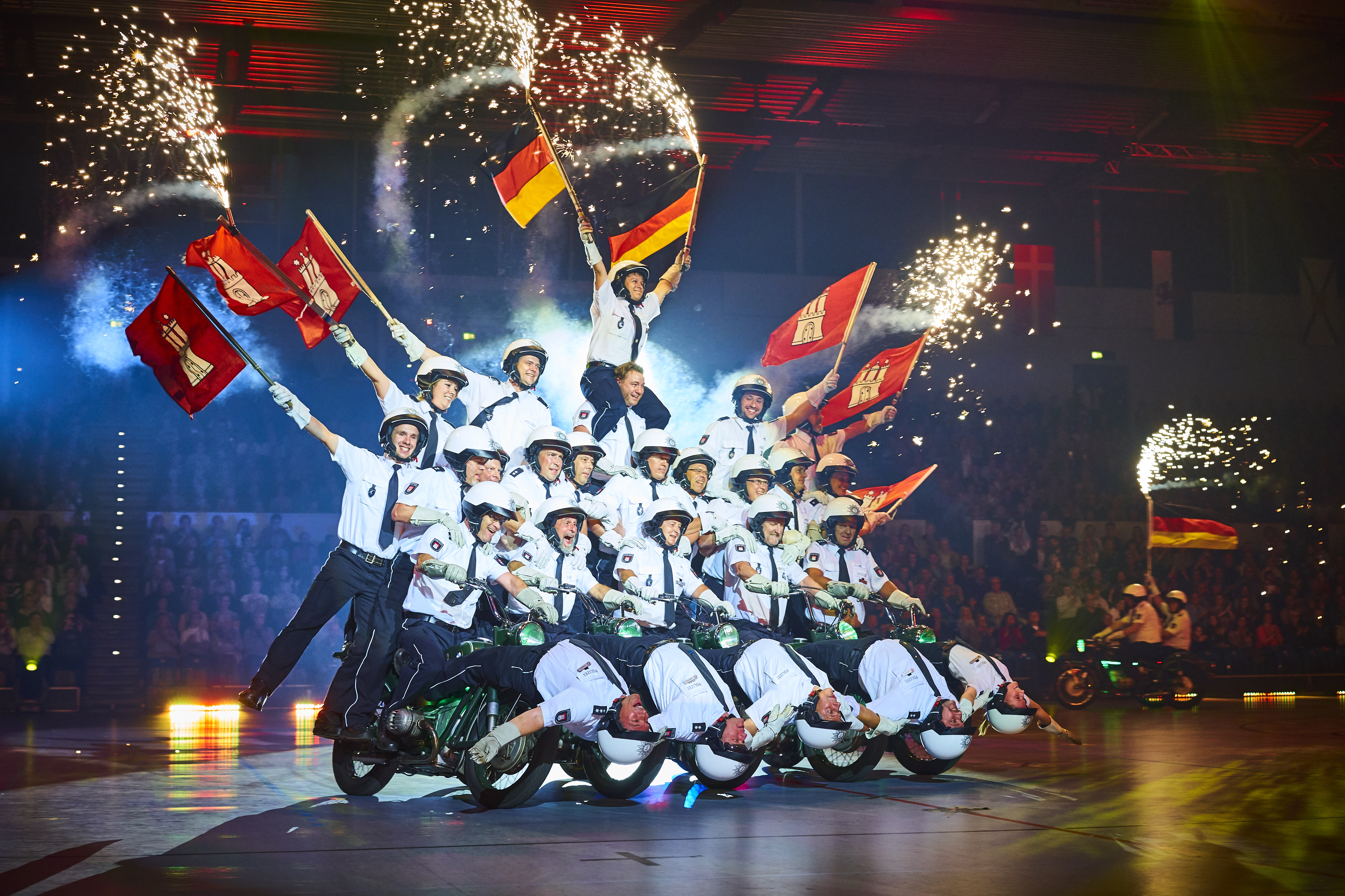 Pyramide mit Polizisten, die unterste Reihe sitzt auf Motorrädern, die Oberen schwenken Fahnen, Feuerwerk im Hintergrund